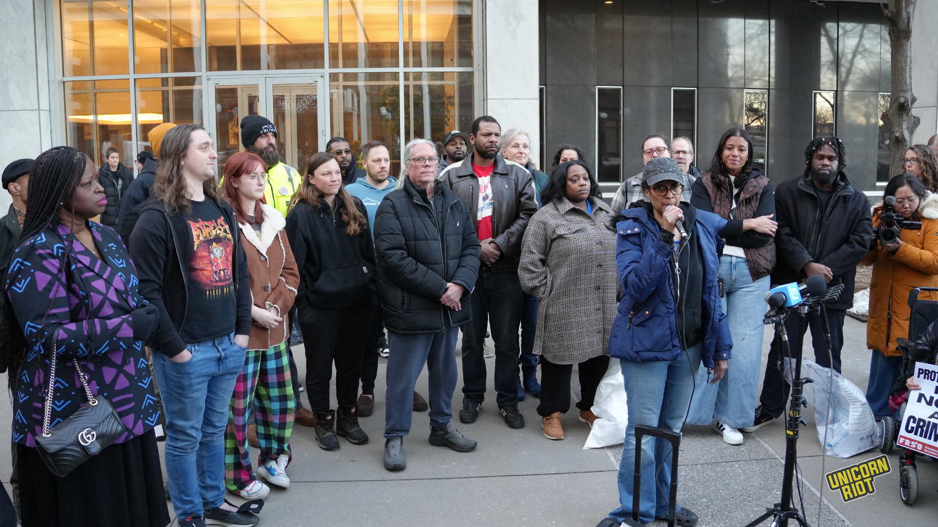A diverse group of people gathered outdoors, some listening intently while one individual speaks into a microphone. In the background, a banner can be seen promoting a message related to activism.