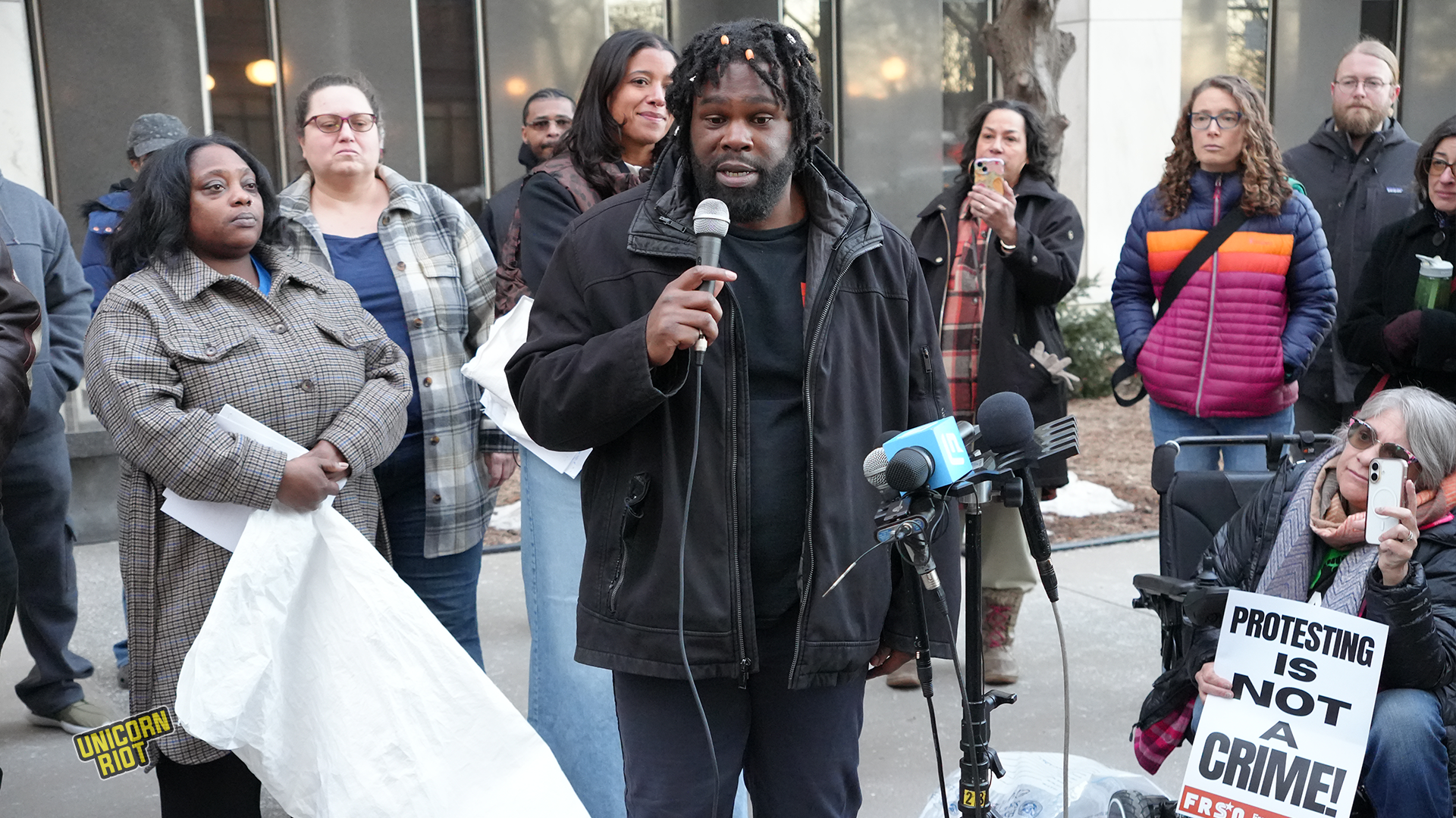 A speaker addresses a crowd at a protest, holding a microphone and delivering a message about civil rights, while supporters listen attentively, some holding signs.